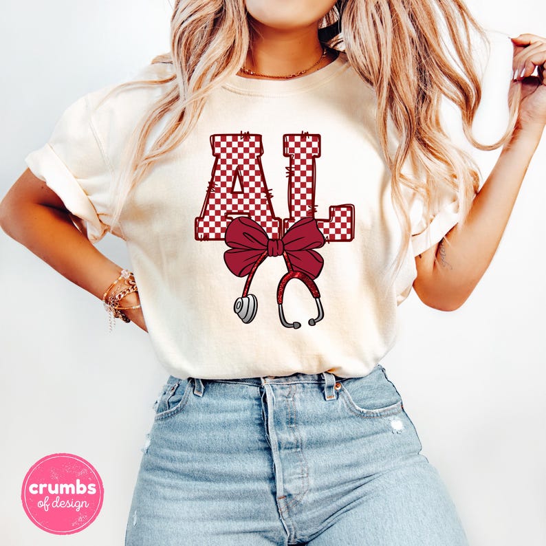 May include: Cream-colored t-shirt featuring the letters "AL" in a maroon and white checkered pattern, with a maroon bow and a stethoscope graphic. The shirt is paired with blue jeans.