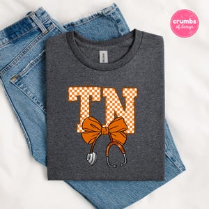 May include: A dark gray t-shirt with the letters "TN" in orange and white checkered pattern, a large orange bow, and a stethoscope design. The shirt is folded on top of blue jeans.