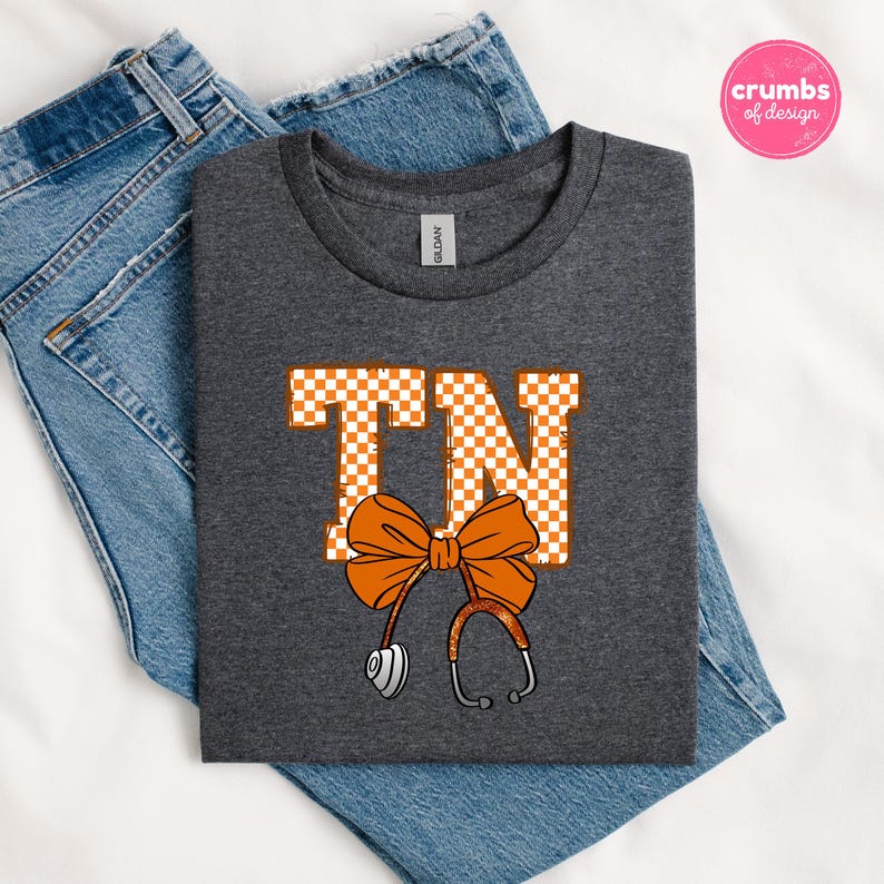 May include: A dark gray t-shirt with the letters "TN" in orange and white checkered pattern, a large orange bow, and a stethoscope design. The shirt is folded on top of blue jeans.