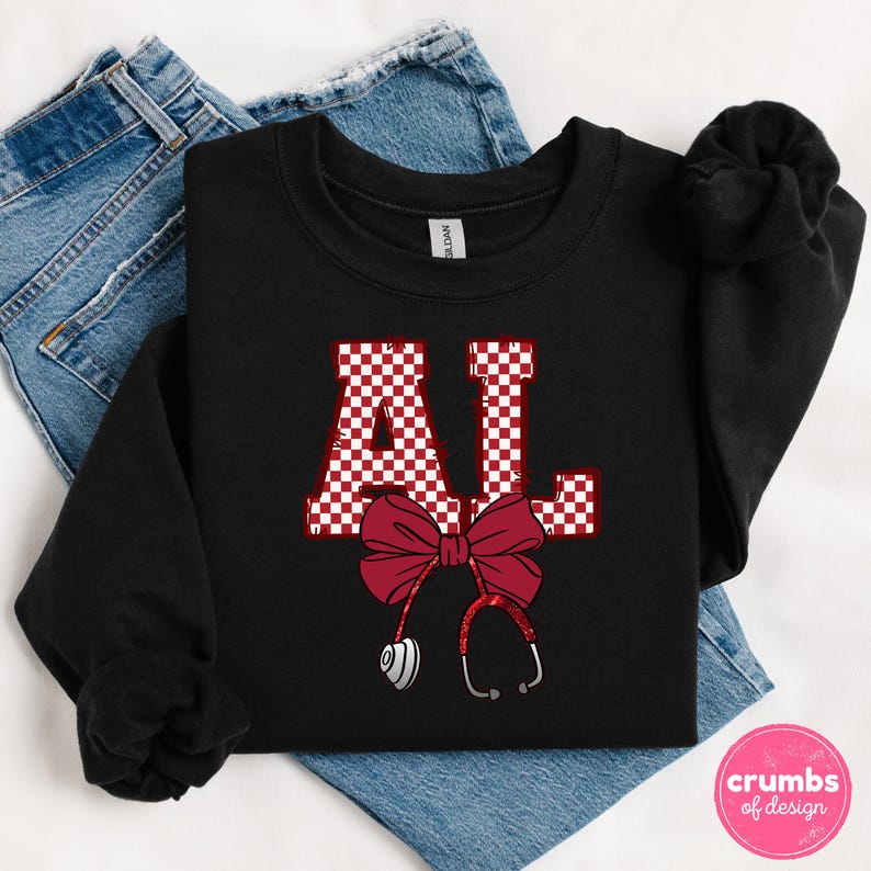 May include: Black sweatshirt with the letters "AL" in a red and white checkered pattern. A red bow and stethoscope design are below the letters. The sweatshirt is folded on top of a pair of blue jeans. The design is from "crumbs of design."