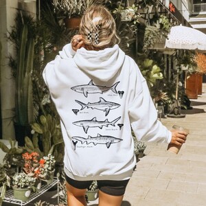 May include: White hoodie with a graphic of four different types of sharks on the back. The hoodie has a drawstring hood and a kangaroo pocket.