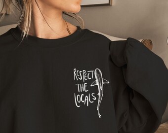 Puede incluir: Sudadera negra con un gráfico blanco de un tiburón y el texto "Respect the Locals".