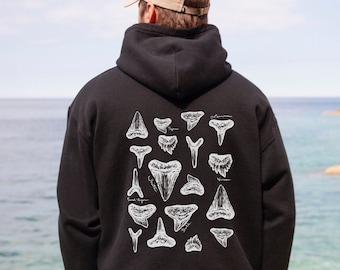 May include: Black hoodie with a white shark tooth design on the back. The hoodie has a hood and a front pocket. The person is wearing a beige baseball cap and beige trousers. The background shows a beach and the sea.