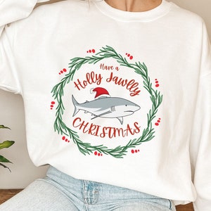 Puede incluir: Sudadera blanca con un dise&ntilde;o de corona de acebo verde con un tibur&oacute;n que lleva un gorro de Pap&aacute; Noel. El texto dice "Have a Holly Jawlly Christmas".