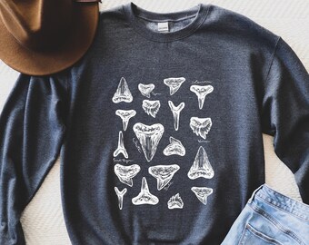 Puede incluir: Una sudadera gris oscuro con un estampado blanco de diferentes dientes de tiburón. La impresión incluye los nombres de las especies de tiburones.