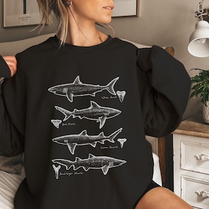 May include: Black sweatshirt with a white graphic of four different types of sharks with their names: White Shark, Bull Shark, Lemon Shark, and Sandtiger Shark.