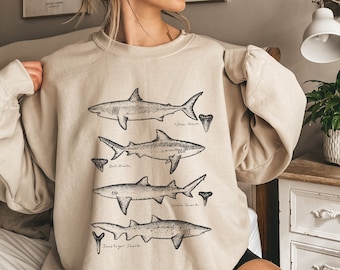 Op de afbeelding: Een beige sweatshirt met een grafische print van vier verschillende soorten haaien en hun tanden. De tekst op het sweatshirt luidt "Walvishaai", "Stierenhaai", "Citroenhaai" en "Zandtijgerhaai".