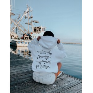 May include: White hoodie with a black hood and a graphic of four different types of sharks on the back. The person is wearing the hoodie and is sitting on a wooden jetty.