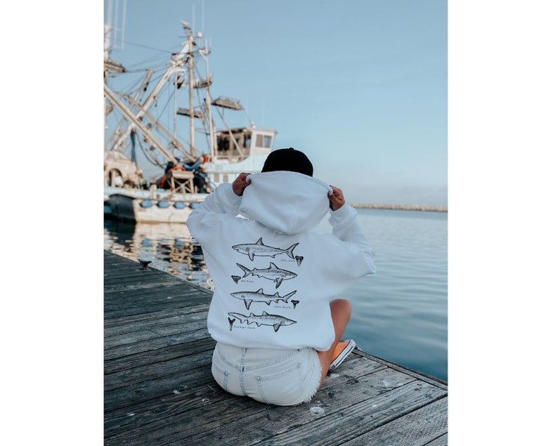 May include: White hoodie with a black hood and a graphic of four different types of sharks on the back. The person is wearing the hoodie and is sitting on a wooden jetty.