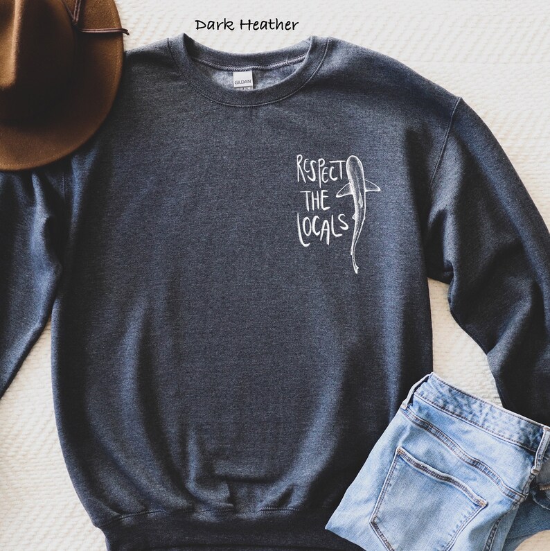 May include: Dark heather grey sweatshirt with a white graphic of a shark and the text "Respect the Locals".
