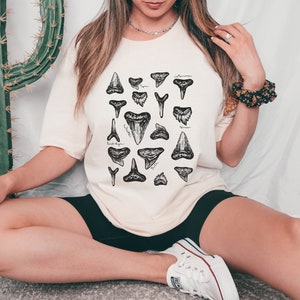 May include: A white t-shirt with a black and white illustration of different shark teeth. The shirt has the names of the shark species written next to each tooth.