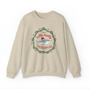 Puede incluir: Una sudadera beige con un gr&aacute;fico navide&ntilde;o que presenta un tibur&oacute;n con un gorro de Pap&aacute; Noel en una corona con el texto "Have a Holly Jawlly Christmas".