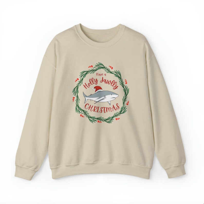 Puede incluir: Una sudadera beige con un gr&aacute;fico navide&ntilde;o que presenta un tibur&oacute;n con un gorro de Pap&aacute; Noel en una corona con el texto "Have a Holly Jawlly Christmas".
