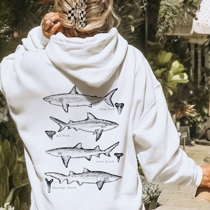 May include: White hoodie featuring a graphic of four different shark species, including a white shark, bull shark, lemon shark, and sand tiger shark. Each shark is drawn in black and white and has a tooth next to it.