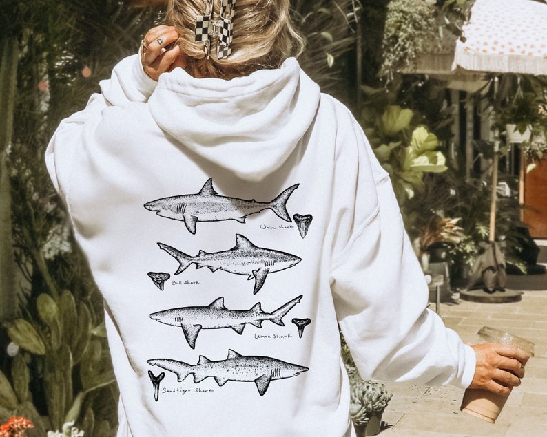 May include: White hoodie featuring a graphic of four different shark species, including a white shark, bull shark, lemon shark, and sand tiger shark. Each shark is drawn in black and white and has a tooth next to it.