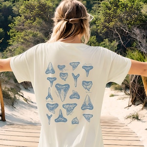 May include: Cream-colored t-shirt featuring a blue illustration of various shark teeth. The design includes different shapes and sizes of teeth, with some labeled. The shirt is worn outdoors.
