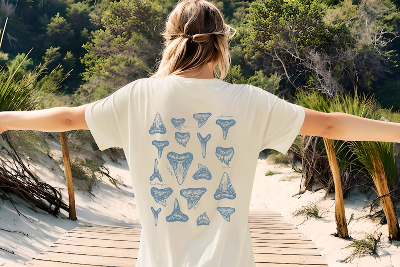 May include: Cream-colored t-shirt featuring a blue illustration of various shark teeth. The design includes different shapes and sizes of teeth, with some labeled. The shirt is worn outdoors.
