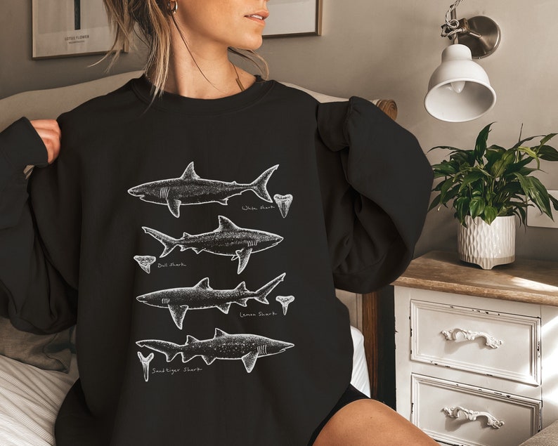 May include: Black sweatshirt with a white graphic of four different types of sharks with their names: White Shark, Bull Shark, Lemon Shark, and Sandtiger Shark.