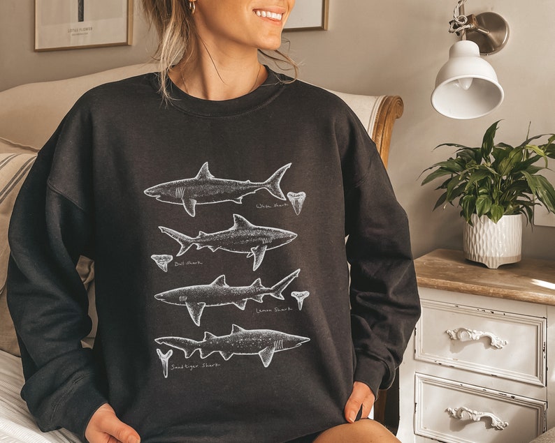 May include: Black sweatshirt with a white graphic of four different types of sharks with their names: White Shark, Bull Shark, Lemon Shark, and Sand Tiger Shark.
