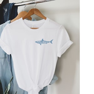 May include: White t-shirt with a light blue shark graphic that reads "Respect Locals." The shirt is hanging on a wooden hanger with other clothing items in the background. The shirt has rolled-up sleeves.