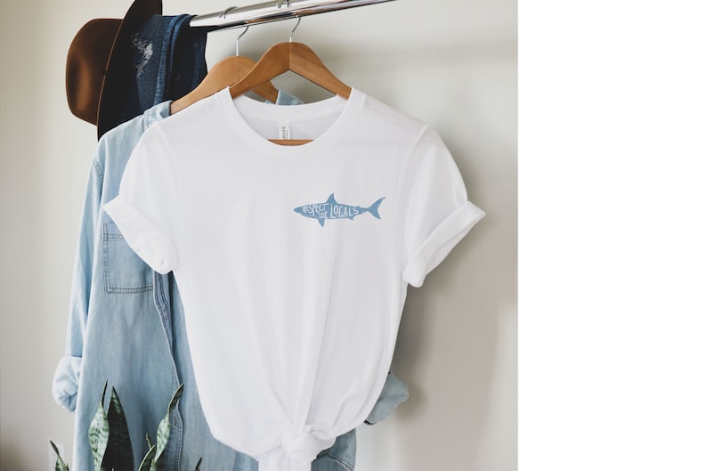 May include: White t-shirt with a light blue shark graphic that reads "Respect Locals." The shirt is hanging on a wooden hanger with other clothing items in the background. The shirt has rolled-up sleeves.