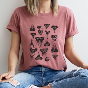 May include: A woman wearing a dusty rose t-shirt with a black and white illustration of various shark teeth. The shirt has a crew neck and short sleeves.