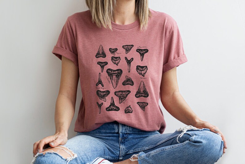 May include: A woman wearing a dusty rose t-shirt with a black and white illustration of various shark teeth. The shirt has a crew neck and short sleeves.