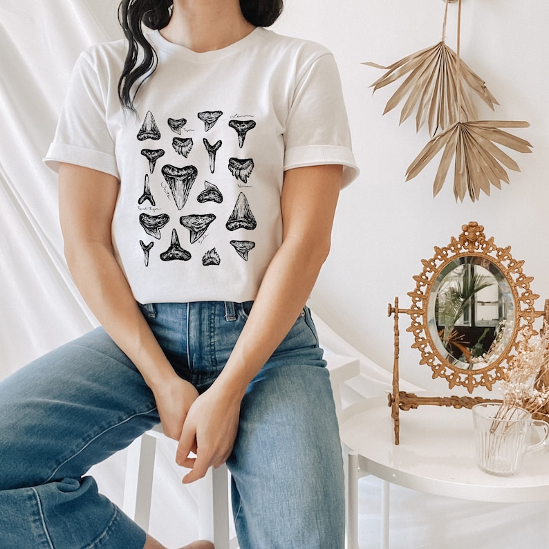 May include: White t-shirt with a black and white illustration of various shark teeth. The shirt is being worn by a person with blue jeans.