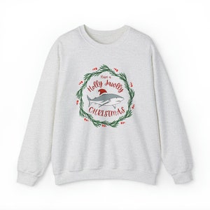 Puede incluir: Una sudadera gris con un gr&aacute;fico navide&ntilde;o que presenta un tibur&oacute;n con un gorro de Pap&aacute; Noel en una corona con el texto "Have a Holly Jawlly Christmas".