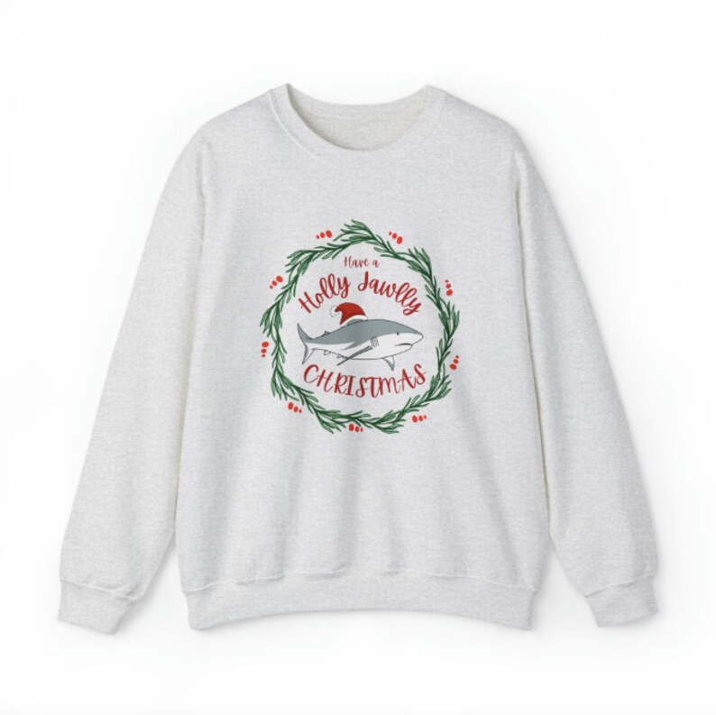 Puede incluir: Una sudadera gris con un gr&aacute;fico navide&ntilde;o que presenta un tibur&oacute;n con un gorro de Pap&aacute; Noel en una corona con el texto "Have a Holly Jawlly Christmas".