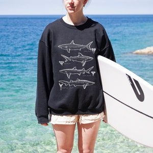 May include: A person wearing a black sweatshirt with a white graphic of different types of sharks. The person is holding a white surfboard with a black stripe and a black letter U on the side.