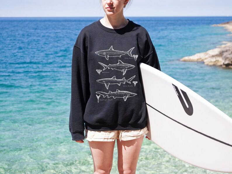 May include: A person wearing a black sweatshirt with a white graphic of different types of sharks. The person is holding a white surfboard with a black stripe and a black letter U on the side.