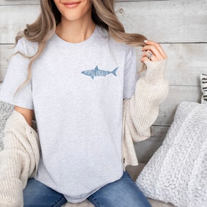 May include: Light gray t-shirt with a shark graphic and the words "Asheville Locals" in blue. The shirt is made of a soft material and is suitable for casual wear. The shirt is a crew neck style.