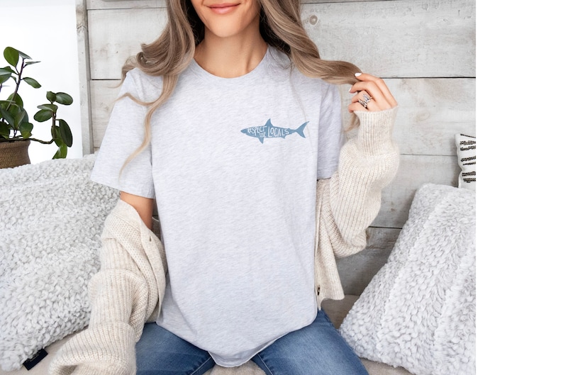 May include: Light gray t-shirt with a shark graphic and the words "Asheville Locals" in blue. The shirt is made of a soft material and is suitable for casual wear. The shirt is a crew neck style.