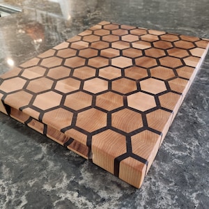 May include: A wooden chopping board with a honeycomb pattern in black and natural wood. The board is made of maple wood and has a natural finish.