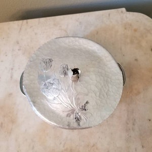 May include: A silver serving dish with a hammered finish and a floral design. The dish has a lid with a knob in the center.