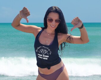 May include: A woman wearing a navy blue tank top and purple bikini bottoms stands on a beach with the ocean in the background. The tank top has the text "Brilliant Mockup Pix YOUR DESIGN GOES HERE". She is wearing sunglasses and giving a thumbs down.
