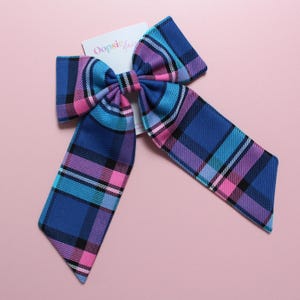 May include: A fabric hair bow featuring a plaid pattern in shades of blue, purple, and bright pink. The bow has two long, pointed tails and is attached to a white display card with the text Oopsie Daisy printed in pink and black lettering.