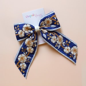 May include: A navy blue hair bow with detailed white and gold floral embroidery. The bow has a white centre knot and is decorated with small gold sequins. The tag reads "Oopsie Daisy".