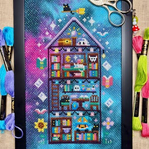 May include: Framed cross-stitch art depicting a whimsical house with multiple rooms filled with books and decorative objects. The artwork features a starry night sky background with a black cat on a broom. Embroidery floss and scissors are included.