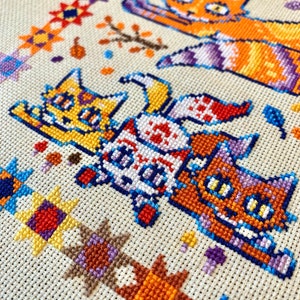 Puede incluir: Bordado de punto de cruz con cuatro gatos de pelaje naranja, amarillo, blanco y marr&oacute;n. Los gatos est&aacute;n dispuestos en fila, con el gato de arriba mirando hacia la derecha y los otros tres mirando hacia la izquierda. El fondo es una tela beige con un borde colorido de formas geom&eacute;tricas.