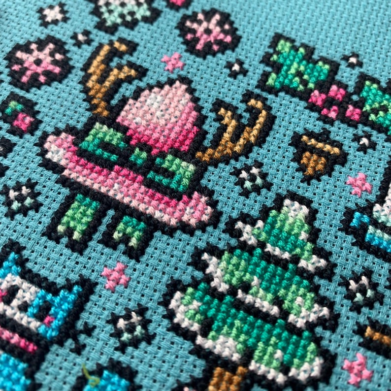 Pu&ograve; includere: Ricamo a punto croce su tessuto azzurro chiaro con un design festivo natalizio. Il disegno include una renna, un albero di Natale e fiocchi di neve, tutti realizzati con filo rosa, verde, marrone e bianco con contorni neri.