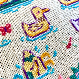 May include: Close-up of a cross-stitch pattern featuring a yellow duck with purple and blue accents. The duck is swimming in a blue and white wave pattern. The background is a white fabric with a grid pattern.