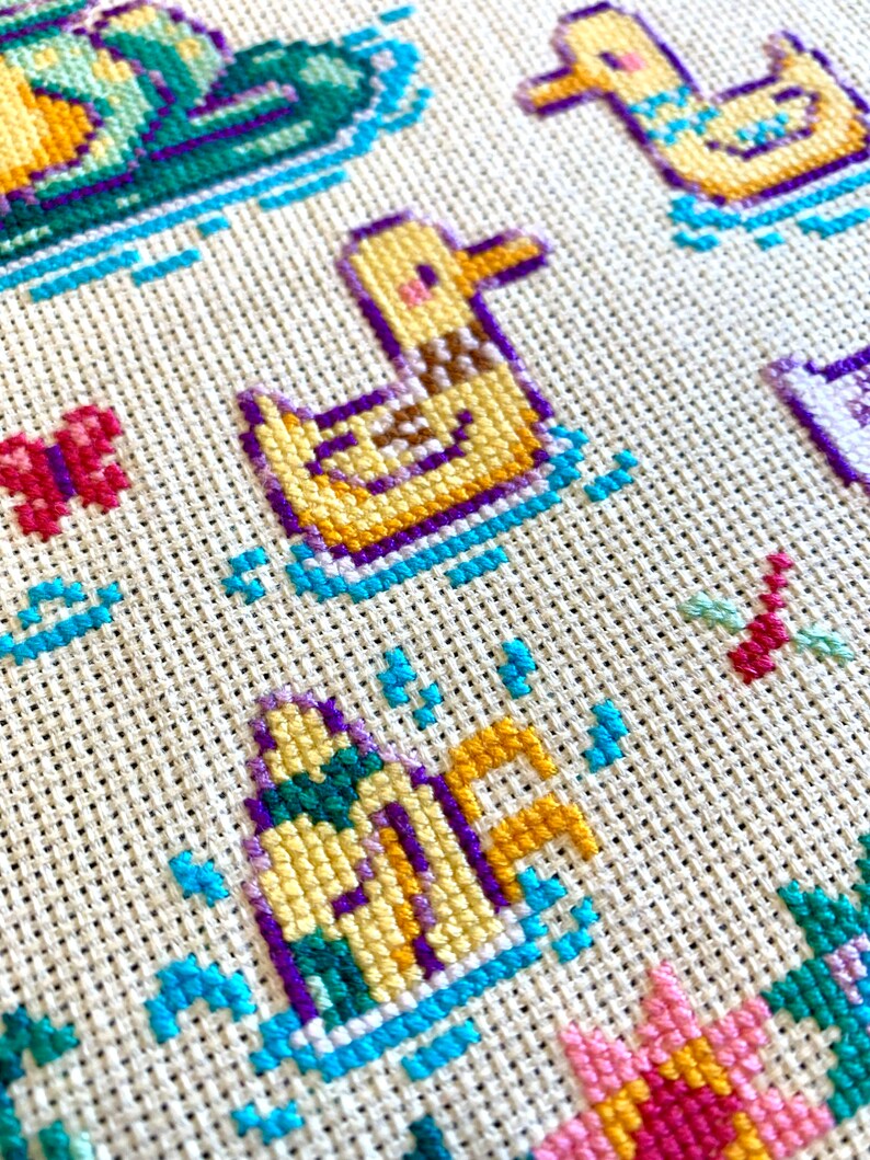 May include: Close-up of a cross-stitch pattern featuring a yellow duck with purple and blue accents. The duck is swimming in a blue and white wave pattern. The background is a white fabric with a grid pattern.