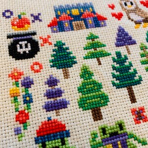 K&ouml;nnte beinhalten: Kreuzstich-Stickerei mit einer fantasievollen Halloween-Szene mit einem lila Haus, einem Kessel, einem K&uuml;rbis, einem gr&uuml;nen Frosch, einer lila Eule und verschiedenen B&auml;umen und Blumen.