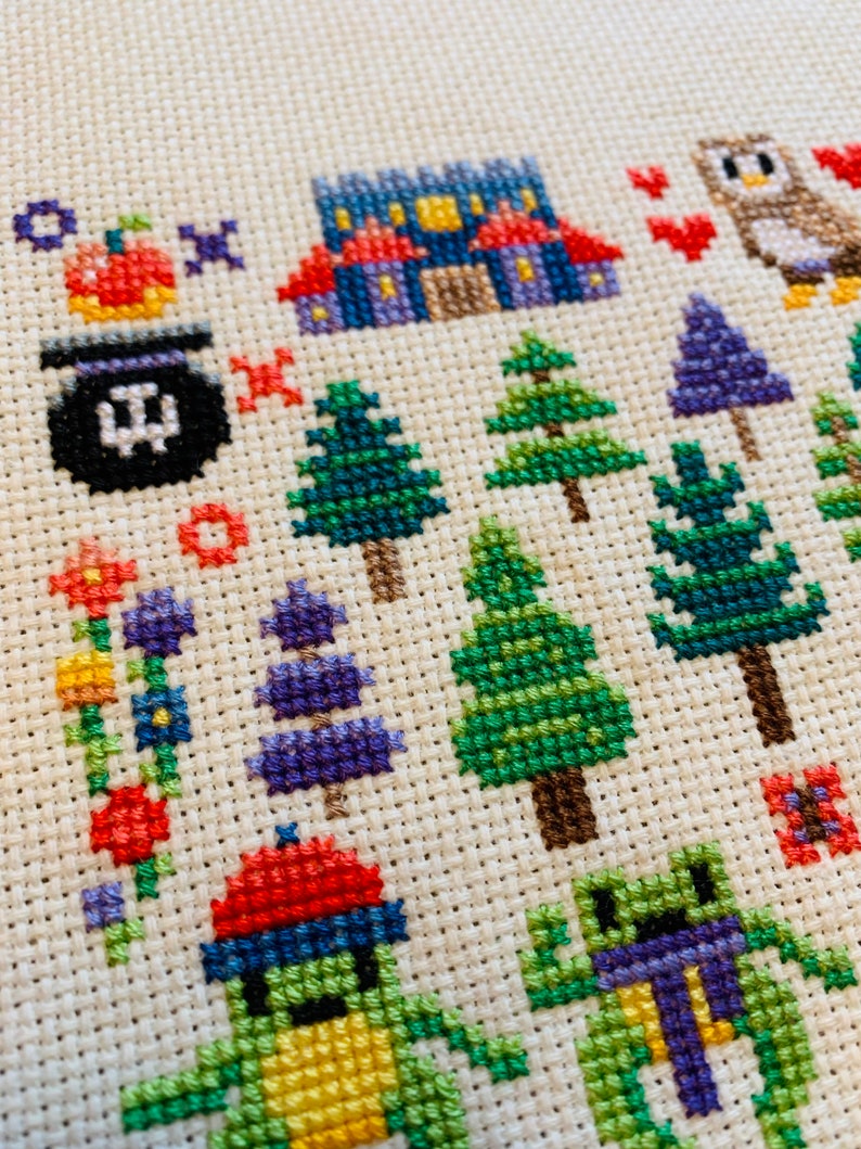 K&ouml;nnte beinhalten: Kreuzstich-Stickerei mit einer fantasievollen Halloween-Szene mit einem lila Haus, einem Kessel, einem K&uuml;rbis, einem gr&uuml;nen Frosch, einer lila Eule und verschiedenen B&auml;umen und Blumen.