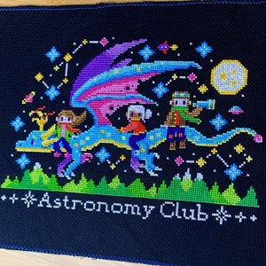 Pode incluir: Bordado de ponto cruz de um drag&atilde;o azul voando por um c&eacute;u noturno estrelado com tr&ecirc;s figuras montadas em suas costas. O drag&atilde;o tem asas rosa, roxas e azuis. As figuras usam roupas coloridas e seguram telesc&oacute;pios. O texto "Astronomy Club" est&aacute; bordado abaixo do drag&atilde;o.