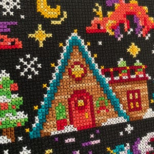 May include: A cross-stitch pattern featuring a gingerbread house with a red door, a green roof, and a yellow star on the front. The house is surrounded by snowflakes, stars, and a crescent moon. There are also two trees and a unicorn in the scene.
