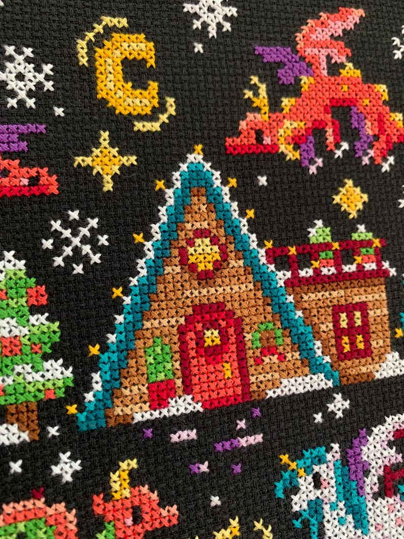 May include: A cross-stitch pattern featuring a gingerbread house with a red door, a green roof, and a yellow star on the front. The house is surrounded by snowflakes, stars, and a crescent moon. There are also two trees and a unicorn in the scene.