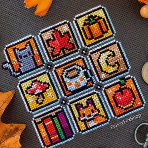 May include: A cross-stitch piece with nine square panels depicting autumn symbols. The panels show a cat, maple leaf, pumpkin, crescent moon, mushroom, coffee cup, apple, books, and a fox. The piece is set against a grey background.